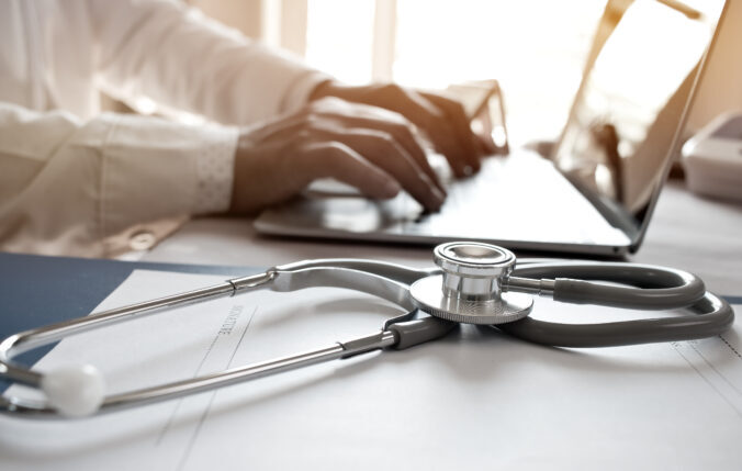 Doctor yn teipio ar liniadur gyda stetosgop ar ochr y gliniadur