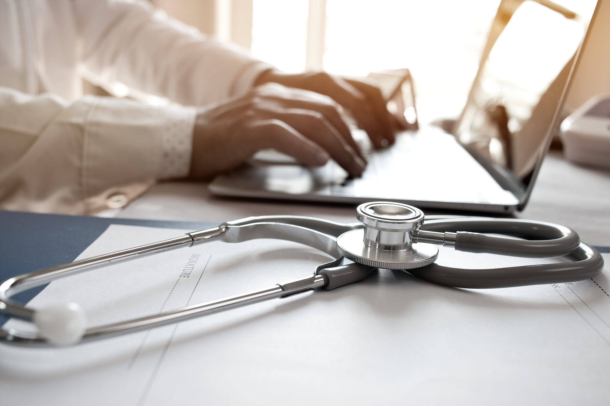 Doctor yn teipio ar liniadur gyda stetosgop ar ochr y gliniadur